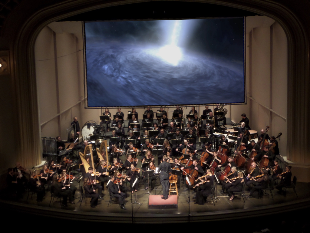 Boulder Phil plays music about stars and astronomers and planets in front of screen displaying beautiful space images